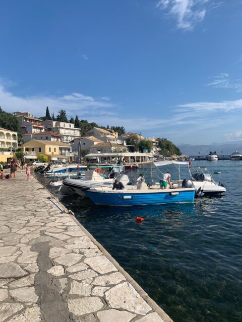 A stone paved walkway next to a harbour where small fishing boats are moored.