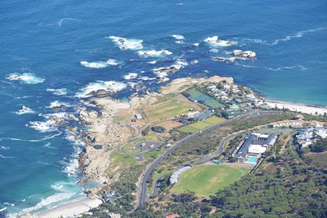 View from the Top of Table Mountain include waves crashing against large rocks and cliffs and green areas with a road running between.