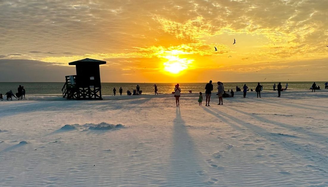 One of the best things to do in Sarasota is to enjoy relaxing on Siesta Key beach at sunset. People are enjoying the orange glow of the sun sinking.