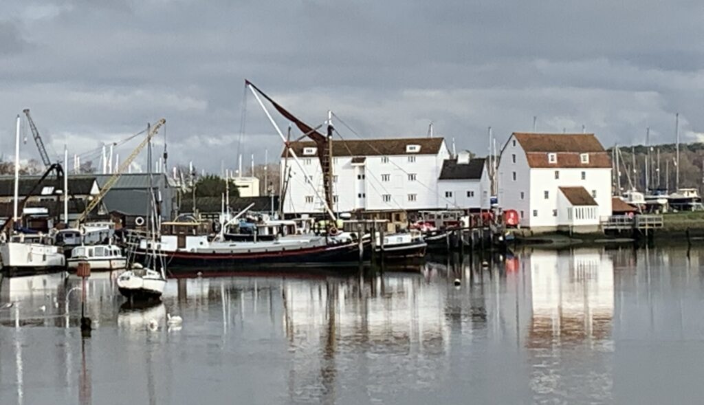 Riverside quay in Woodbridge where house boats and fishing vessels are moored. Old mill with whitewashed cascade sits at the riverside edge. Cranes are located to lift boats in and out of the water.