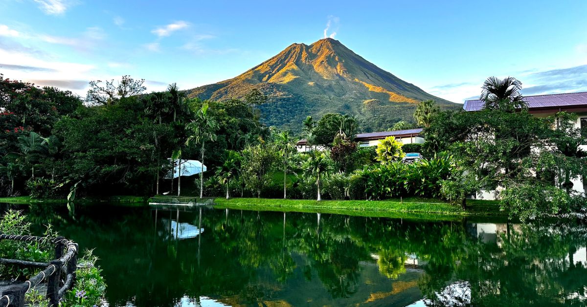 Arenal Volcano with  light puffs of smoke coming from the top. In front of volcano is a lake where volcano is reflected in.