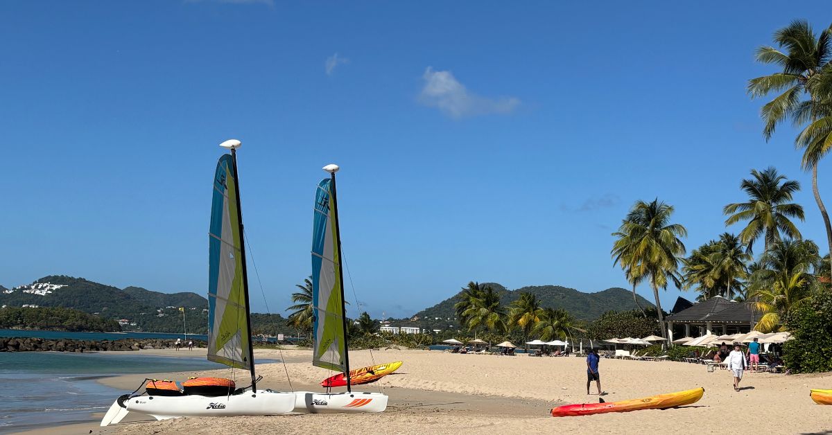 Sandy beach with kayaks on the sand and two small boats pulled with sails on them. Palm trees and sun loungers dotted around the edge.